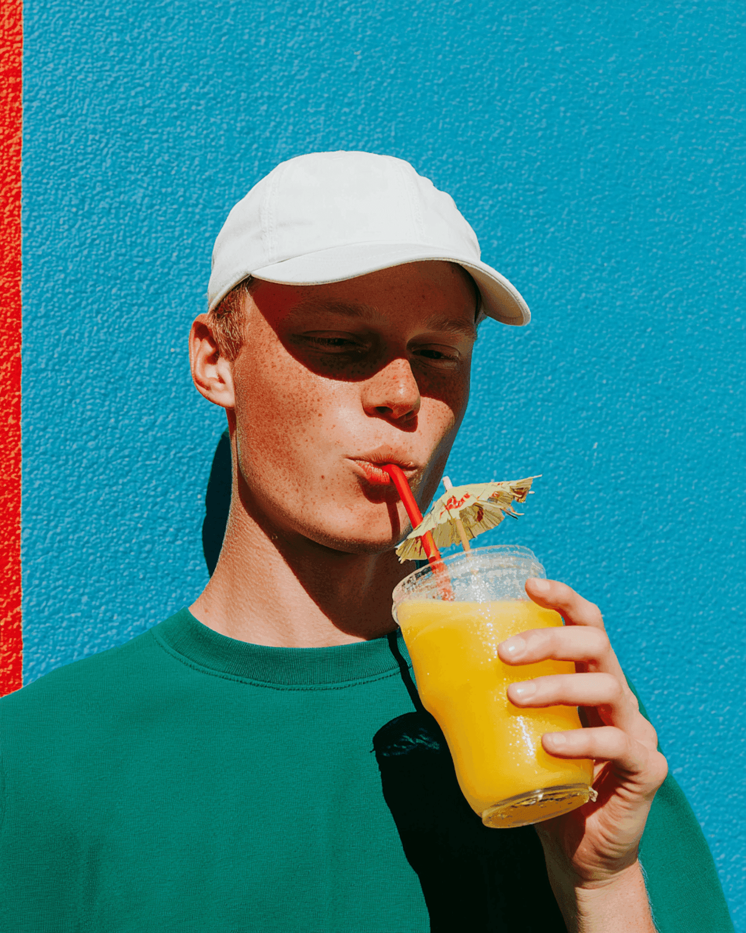Young man in a white cap sipping orange juice against a blue wall